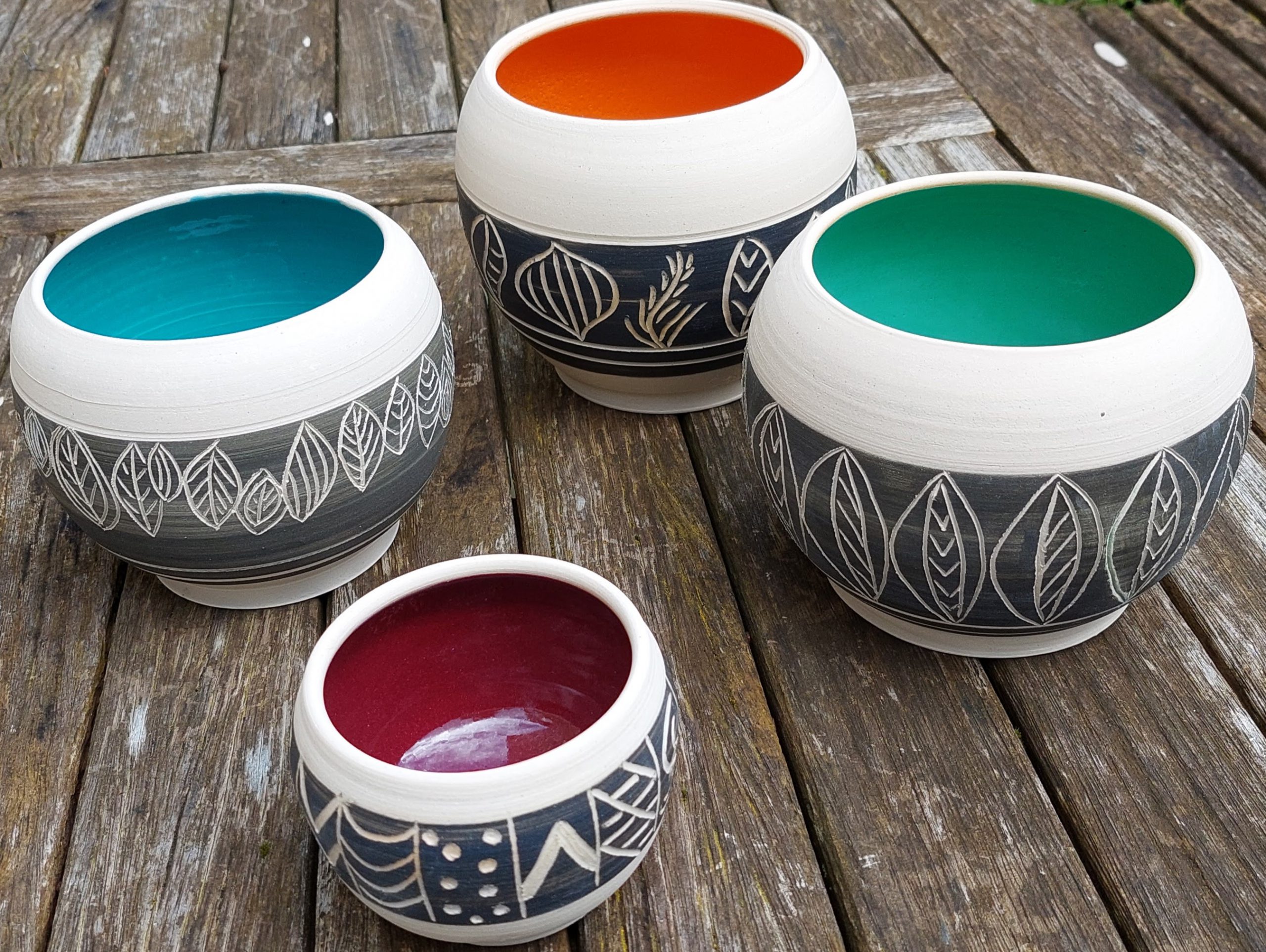 Four stoneware wheel thrown bowls with sgraffito patterns on charcoal on the outides and glazed insides in blue, orange, green and red, respectively.
