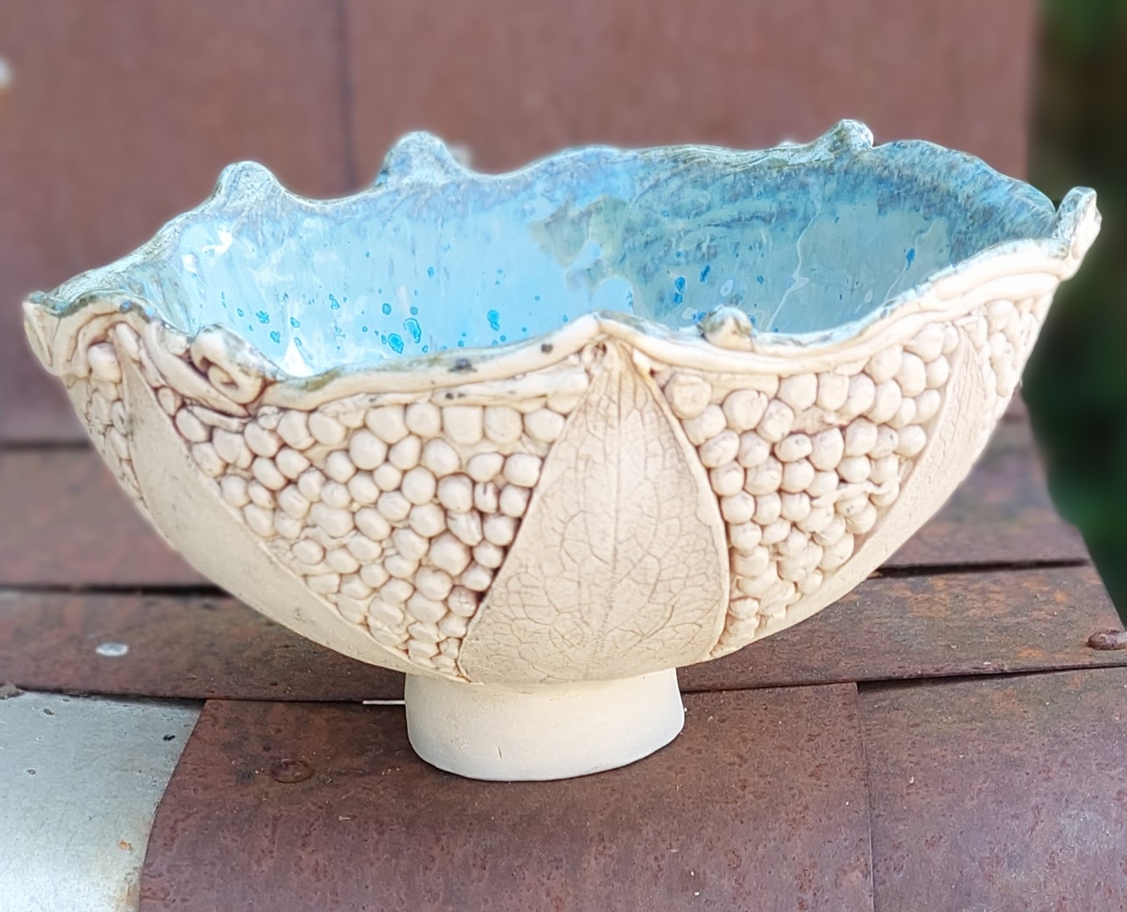Hand thrown bowl on a stem. Pale blue inside. Coils and leaves outside in natural clay.